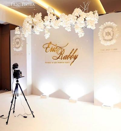 ฉากหลังงานแต่งงานสีขาวสะอาดตา ตกแต่งด้วยตัวอักษรชื่อบ่าวสาวสีทองนูนสวยงาม ด้านบนประดับพวงดอกกล้วยไม้สีขาวขนาดใหญ่ห้อยระย้าดูมีมิติ พร้อมการจัดแสงไฟอัพไลท์จากพื้นเพื่อสร้างความสว่างและหรูหรา