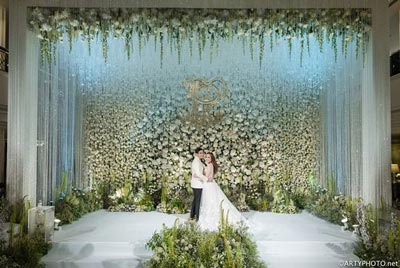 ฉากหลังงานแต่งงานผนังสีขาว ตกแต่งด้วยชื่อบ่าวสาวสีทอง ประดับพุ่มใบไม้สีเขียวชอุ่มและดอกไม้สีขาวที่มุมฉาก พร้อมแจกันดอกไม้ทรงสูงและพุ่มดอกไม้ที่พื้นหญ้าเทียม สร้างบรรยากาศสดชื่นและเรียบง่าย
