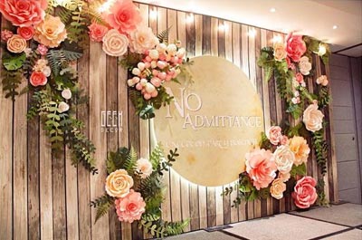ฉากแบ็คดรอปงานแต่งงานทำจากไม้ระแนงสีน้ำตาลคลาสสิก ตกแต่งด้วยดอกไม้กระดาษขนาดใหญ่โทนสีส้มพาสเทลและใบเฟิร์นสีเขียว พร้อมป้ายวงกลมไฟซ่อนระบุข้อความกลางฉาก