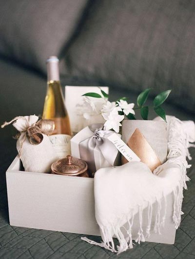 กล่องของขวัญสีขาวทรงสี่เหลี่ยมจัดเซต Wedding Welcome Gift ภายในมีขวดไวน์ขาว, กล่องของขวัญผูกโบว์สีเงินพร้อมป้าย "Welcome", ถุงผ้าขนาดเล็กประดับกระดิ่งคู่, ตลับโลหะสีทองแดง และกระถางเซรามิกสีเทาคาดทอง ตกแต่งด้วยดอกไม้สีขาวและผ้าคลุมไหล่สีขาวมีพู่