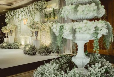 ฉากหลังงานแต่งงานสีขาวขนาดใหญ่ประดับดอกไม้สดสีขาวและใบไม้ประดิษฐ์อัดแน่นเต็มพื้นที่ ตกแต่งด้วยแจกันทรงสูงสีเงินวางบนแท่นสีขาว พร้อมเลเยอร์พุ่มดอกไม้ด้านหน้าที่ดูมีมิติและหรูหรา