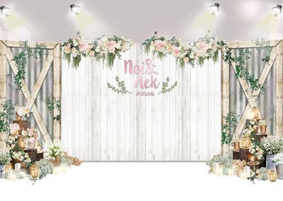 ฉากหลังงานแต่งงานผนังไม้ระแนงสีขาวประดับชื่อบ่าวสาวสีชมพูและดอกไม้สด ขนาบข้างด้วยโครงไม้รูปกากบาทและผนังสังกะสี ตกแต่งเพิ่มเติมด้วยพุ่มดอกไม้สีขาว เทียนหอม และไฟประดับวอร์มไวท์