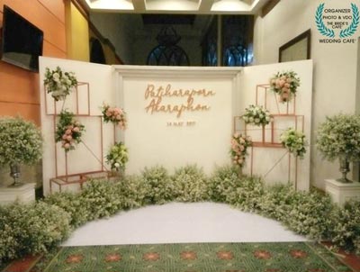 ฉากหลังงานแต่งงานสีขาว ตกแต่งตรงกลางด้วยชื่อบ่าวสาวสีทอง ประดับด้วยโครงเหล็กทรงสี่เหลี่ยมสีโรสโกลด์ที่มีช่อดอกไม้สีชมพูขาววางสลับไปมา พร้อมพุ่มดอกคัตเตอร์สีขาวหนาแน่นที่พื้นด้านล่าง