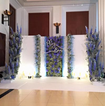 ฉากหลังงานแต่งงานสีขาว ตกแต่งตรงกลางด้วยผนังดอกไม้สีม่วงน้ำเงินไล่เฉดสีสวยงาม ประดับชื่อบ่าวสาวสีขาวนูน มีเลเยอร์พุ่มดอกไม้ทรงสูงสีฟ้าม่วงขนาบข้าง พร้อมการจัดแสงไฟอัพไลท์สร้างบรรยากาศหรูหรา