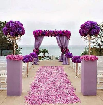 ทางเดินงานแต่งงานตกแต่งด้วยโทนสีม่วงและชมพูเข้ม มีซุ้มม่านสีม่วงประดับดอกไม้ และพุ่มดอกไม้ทรงกลมบนเสาสูงขนาบสองข้างทางเดินที่เต็มไปด้วยกลีบดอกไม้สีชมพู